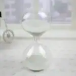 Sablier en verre avec sable blanc, symbole du temps qui passe, posé sur une surface claire avec un décor minimaliste en arrière-plan.