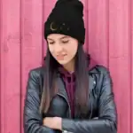 Jeune femme portant un bonnet noir avec motif lunaire et étoiles, vêtue d'une veste en cuir noire et d'un sweat à capuche violet, posant devant un mur en bois rose.