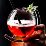 Cocktail coloré dans un verre en verre transparent avec garniture verte, présenté sur un fond noir.