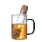 Tasse en verre avec infuseur à thé, remplie d'eau chaude infusant des feuilles de thé, avec bouchon en liège posé sur le dessus.