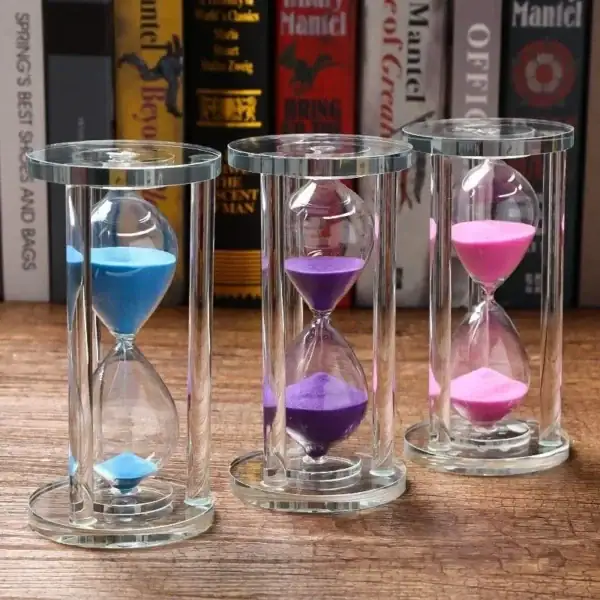 Sablier coloré en verre avec trois tubes contenant du sable bleu, violet et rose, placé sur une surface en bois avec des livres en arrière-plan.
