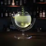 Cocktail lumineux dans un bocal scientifique avec une plante verte flottante, présenté sur un support en verre, ambiance de bar avec étagères de bouteilles en arrière-plan.