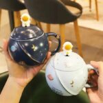 Tasse cosmique avec couvercle et figurine astronaute, décorée de motifs planètes, étoiles et lune, idéale pour les amateurs d'astronomie et de décoration scientifique.