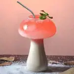 Verre en forme de champignon contenant un cocktail rose avec une branche de menthe, décoratif et original.