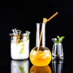 Vase de laboratoire contenant une boisson orange avec une paille, accompagné d'autres accessoires de laboratoire en verre, sur fond noir.