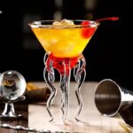 Verre à cocktail scientifique avec décor en forme de tentacules en verre transparent, rempli d'un cocktail jaune-orange avec cerise, ambiance de bar moderne.