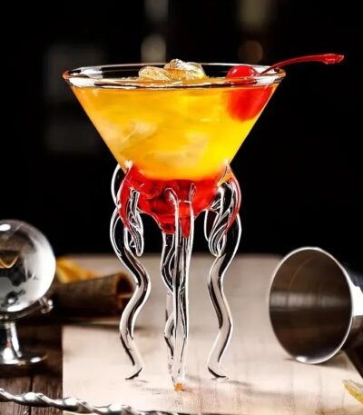 Verre à cocktail scientifique avec décor en forme de tentacules en verre transparent, rempli d'un cocktail jaune-orange avec cerise, ambiance de bar moderne.