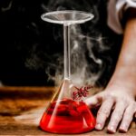 Flacon de laboratoire en verre contenant un liquide rouge fumant, posé sur une surface en bois, avec une main à côté, illustrant une expérience scientifique.