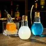 Trois ampoules décoratives remplies de liquides colorés (orange, blanc, bleu) sur une table en bois, ambiance chaleureuse et moderne.