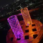 Deux vases en cristal lumineux, l'un violet et l'autre orange, posés sur une table en bois avec vue sur la ville illuminée la nuit.