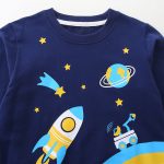 Vêtement pour enfant avec motif spatial, comprenant une fusée, des étoiles, une planète et un télescope, parfait pour les amateurs d'astronomie et d'exploration spatiale.