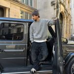 Homme en tenue décontractée posant devant une voiture noire dans une rue urbaine.
